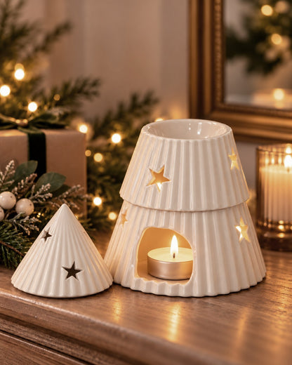 Cream-coloured ceramic Christmas tree-shaped oil burner wax warmer with star cutouts, displayed on a wooden mantelpiece with warm holiday lights in the background.