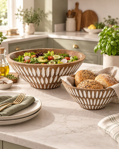 Two decorative mango wood bowls on a marble kitchen counter, one filled with a colourful salad and one with bread rolls, with plates and greenery in a bright kitchen setting.