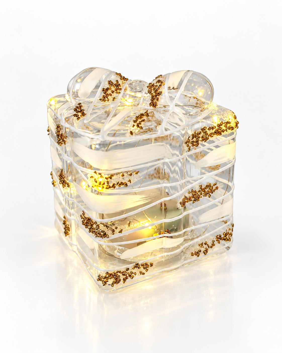 A clear glass gift box sculpture with a bow, illuminated from within with warm yellow lights and decorated with scattered gold glitter