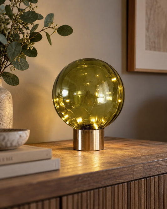 A spherical amber-green glass table lamp with warm yellow LED fairy lights inside, resting on a wooden console table in a styled interior setting.