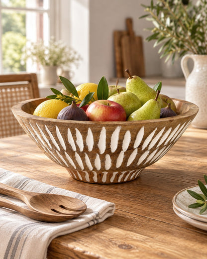 A large round mango wood bowl with a carved geometric pattern sits on a rustic wooden table, filled with fresh fruit including pears, apples, lemons, and figs. A striped napkin with wooden spoons rests in the foreground, and a softly blurred kitchen interior is visible in the background.
